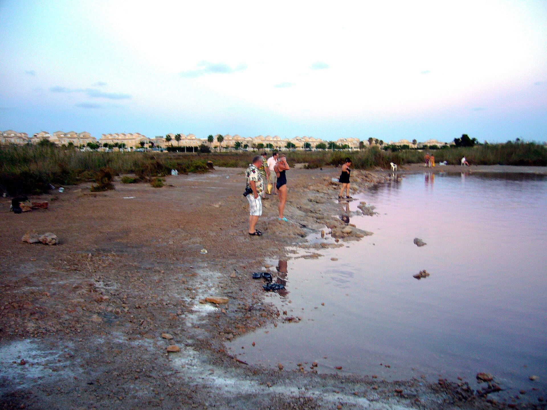 Personas en la orilla de la laguna rosa con urbanización al fondo